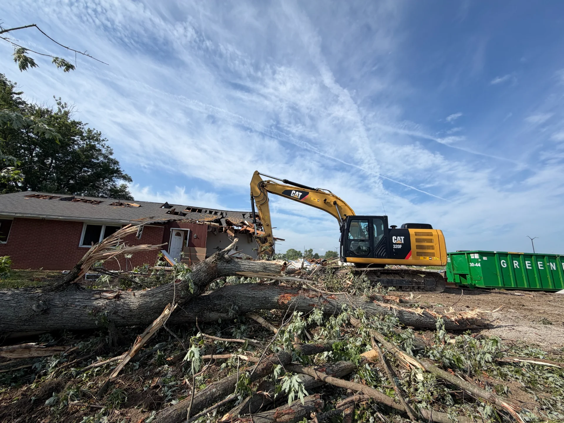Excavator clearing a site with a partially demolished building and felled trees