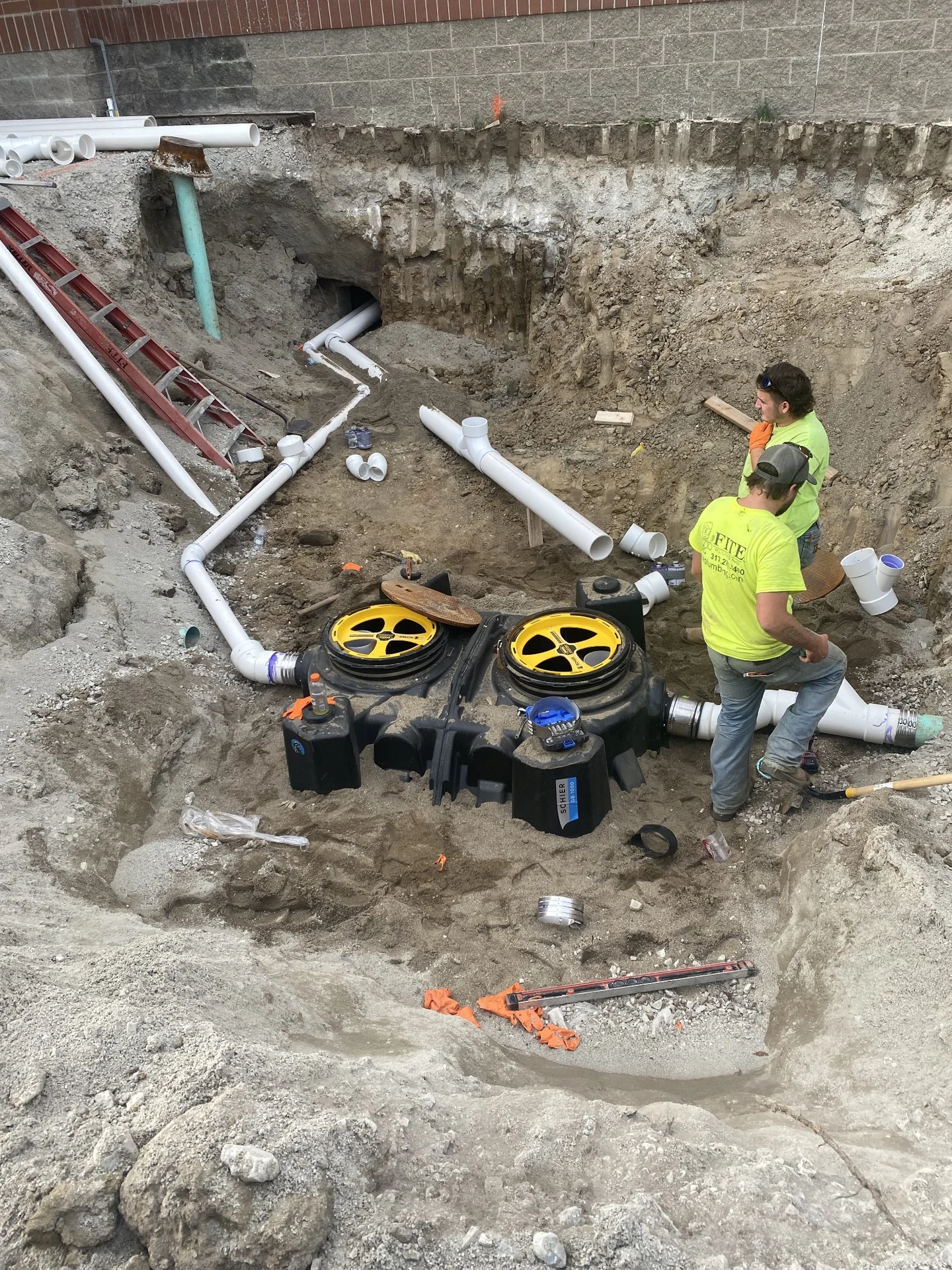 Crew assembling white underground piping and black utility tanks beside a commercial building