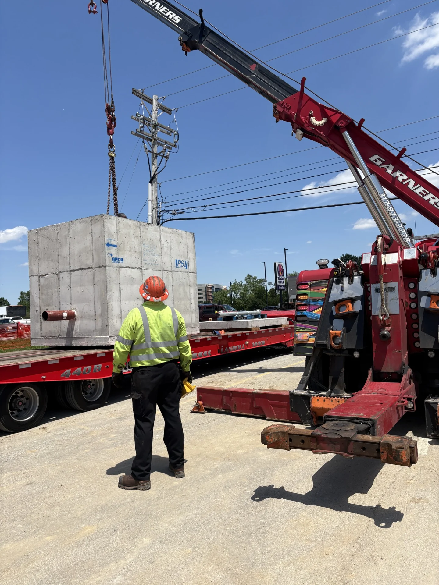 Crane handling a precast concrete utility structure beside a crew member at a commercial jobsite